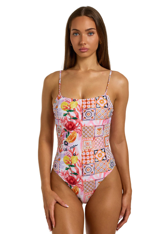 Front view of a woman with dark brown hair wearing the colorful La Mer One Piece swimsuit with thin straps, featuring a vibrant patchwork of floral and geometric patterns, including fruits, flowers, tiles, and the word "la mer" on pink background, non-padded style on a white backdrop, against a plain white backdrop, head to just above the knees visible, right hand resting on right thigh.